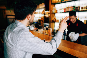 A picture of customer sitting towards the barman. He wants to order some cocktails and showing it with his right hand. THe barman doesn't see it.