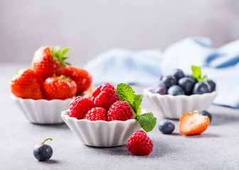 Fresh ripe raspberries, strawberries and bluebetties in white bowls on light gray concrete background. Healthy food concept with copy space.