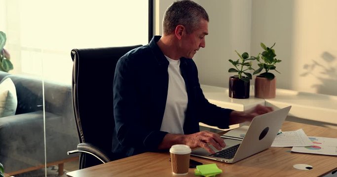 Casually dressed mature businessman focused on his work while sitting at a desk in his office using a laptop