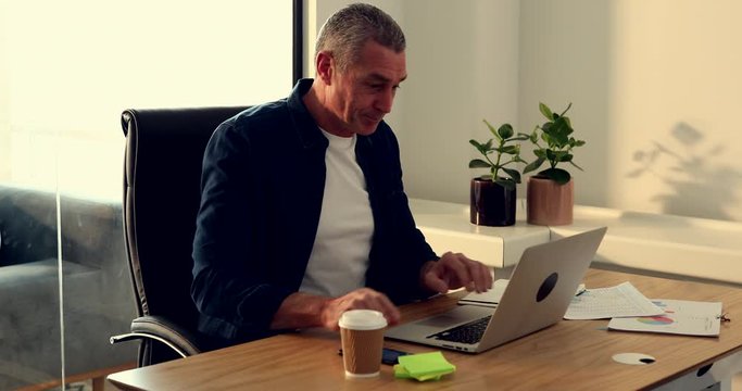 Smiling mature businessman talking on his cellphone while sitting at a desk in his office working online with a laptop