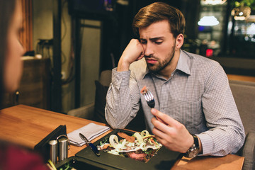 Pictue of guy sitting together with his girlfriend and eating some food they have ordered. Man has found some meat on the plate. He is looking to the piece of meat with suspicious sight. - obrazy, fototapety, plakaty