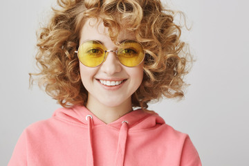 Adorable curly-haired woman with fashionable yellow sunglasses smiling at camera over gray background. Cute artist spends leisure in cafe with friend. Colourful girl ordering food