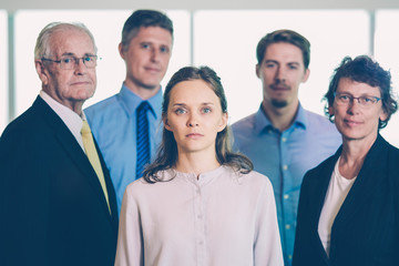Closeup of business team of five serious diverse people standing and looking at camera with big window in background. Two blurred businessmen are in background. Front view.