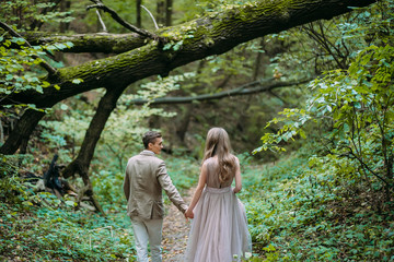 Naklejka na ściany i meble A happy couple is walking on a trail in an autumn forest. Bride and groom are looking at each other on nature.