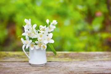 Delicate jasmine flowers in vase on old wooden board, green blurred background