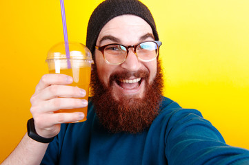 Fototapeta premium Cole up Portrait of attractive young bearded man in hat and glasses standing and drinking orange juice over yellow background and making selfie