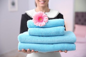 Young maid holding stack of towels and flower in hotel room