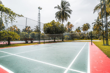 Wide-angle side view of the badminton court located in luxury resort in the Maldives: red and green marked field, palms around, lighting masts, red flower, lanterns and plants; warm sunny day