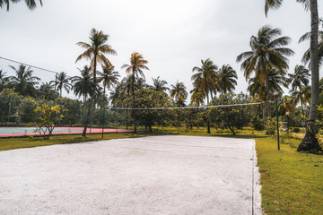 Fototapeta premium Side wide-angle view of the volleyball court: coral sand on the ground, multiple palm trees and other plants around, paled tennis court on the right; bright summer day in a luxury Maldives resort