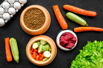 Dry pet food with natural ingredients. Raw meat, vegetables zucchini and carrot near eggs on black backgroud top view pattern