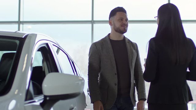 PAN of cheerful bearded man in tweed jacket talking with unrecognizable saleswoman while choosing car at dealership