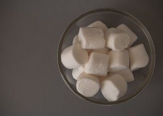 The marshmallows in a clear bowl on the gray background. The white sugar-based candy, sweet, soft, chewy confection made with sugar and gelatin.
