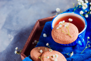 Morning scene with a small blue teacup and oatmeal cookies on a wooden tray with a blue linen napkin. High key snack concept with homemade pastry and spring gypsophila flowers.