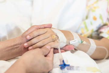 Nursing caretaker concept with child patient (girl kid) sleeping in bed with family caregiver, nurse or parenting mother's hand support in blur medical hospital or clinic background (focus on hand)