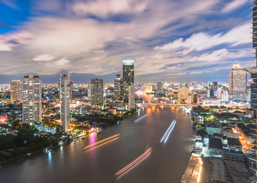 Cityscape night aerial view from rooftop bar over Bangkok's Chao Phraya river CBD and hotel business district after sunset with tourist party sightseeing boat on water travel transportation mode