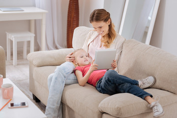 Modern device. Positive joyful blonde girl lying on her mothers legs and playing on a tablet while resting at home