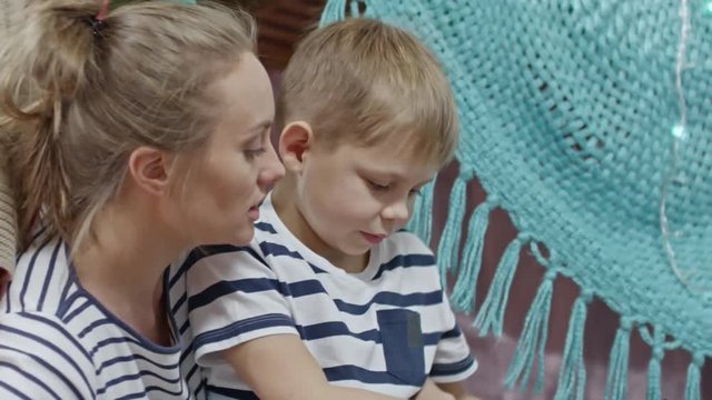 Beautiful woman sitting with cute little son near play tent at home; talking and smiling with boy while he turning on flashlight