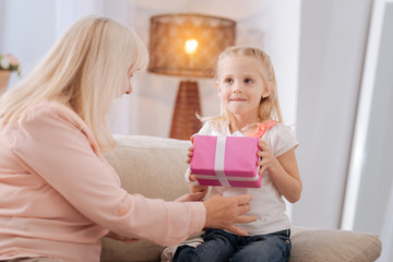 Birthday gift. Joyful nice cute girl smiling and holding a gift box while being together with her grandmother