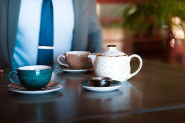 A man in a gray business suit and tie is sitting at the table and pouring tea from the brewer. Lunch break, time out