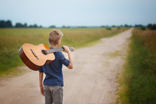 Handsome boy with guitar walking on the road in summer day. Back view