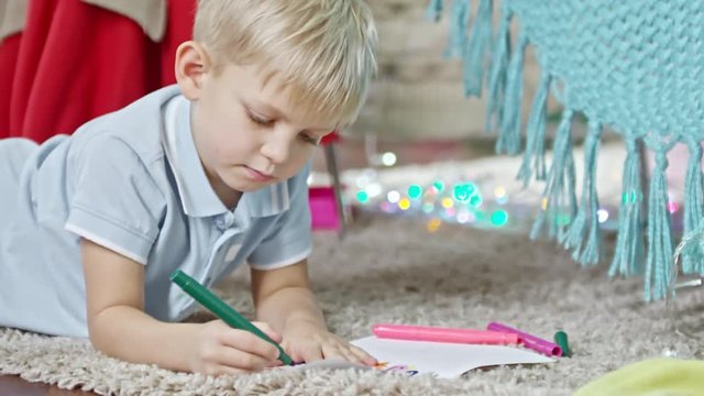 Adorable blonde little boy lying on carpet on the floor and drawing on paper with felt tip pen