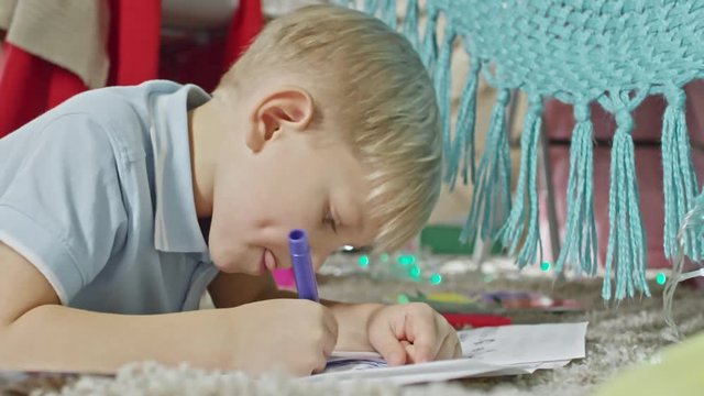 Closeup of concentrated little boy sticking tongue out while lying on carpet at home and drawing picture with felt tip pen