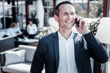 Phone communication. Happy positive nice businessman smiling and holding his smartphone while talking on the phone