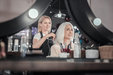 Professional hairstyle. Nice delighted professional hairdresser smiling and straightening hair of her client while looking into the mirror