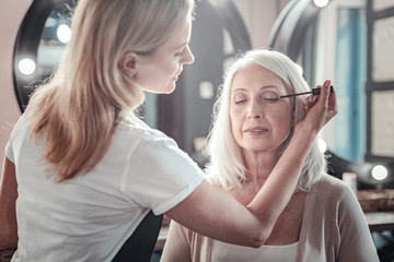 Eye cosmetics. Nice pleasant aged woman sitting on the chair and closing her eyes while having her eyelashes painted