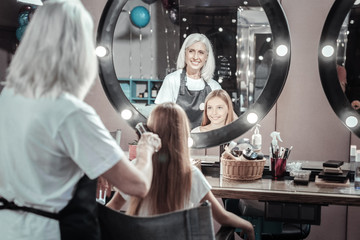 New hairstyle. Nice delighted aged woman smiling and brushing girls hair while working in the beauty salon