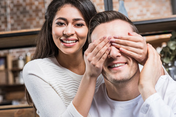 happy young african american woman closing eyes to smiling caucasian boyfriend at home