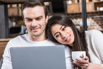 Fototapeta premium smiling multiethnic couple using laptop together while woman drinking tea