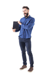 Funny expressive young bearded man presenting blank tablet screen winking at camera. Full body isolated on white background. 