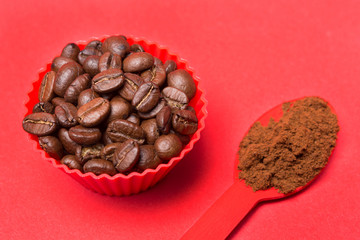 coffee beans in a red container on a red background, next to a red spoon with ground coffee powder