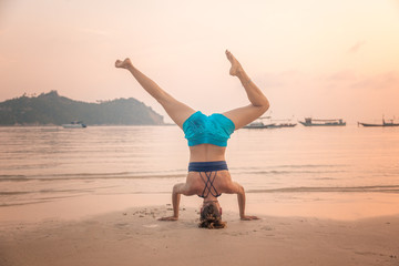 Young woman acrobat stands on her head on a sandy beach on the background of a beautiful sea