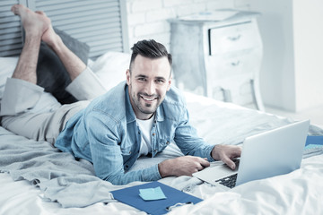 Online education. Cheerful handsome positive man smiling and lying on the bed while doing an online course
