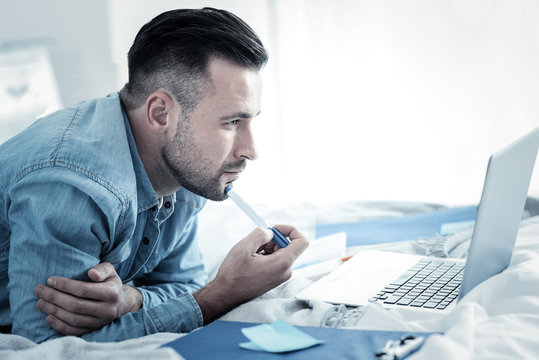 New ideas. Serious handsome smart man holding a pen and looking at the laptop screen while thinking about his project