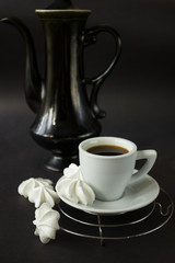 A white cup of espresso on a white saucer with meringues. Black coffee pot. Dark background. serving option