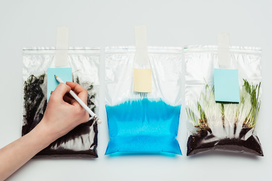 cropped image of woman signing plastic bags with soil, water and seedling isolated on white, earth day concept