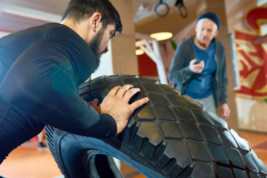 Portrait of bearded Middle-Eastern man flipping tire during training with coach in martial arts club, copy space