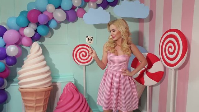 Portrait of a Barbie girl with blond curly hair in a pink dress and a big candy in her hands at a candy party. Barbie style.