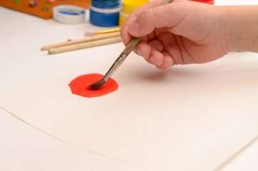 A child's hand draws gouache on white paper with a brush.