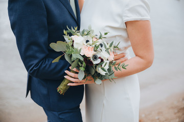 guy in suit and girl in white dress standing on the shore of the lake and holding wedding bouquet of flowers and greens
