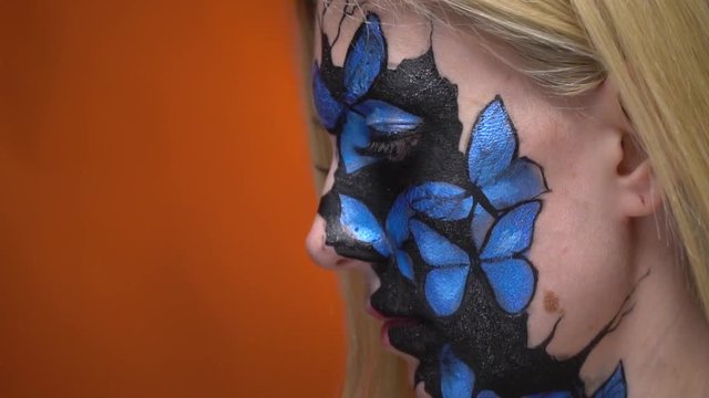 Blonde with body painting on her face in the form of blue butterflies looks at her friend