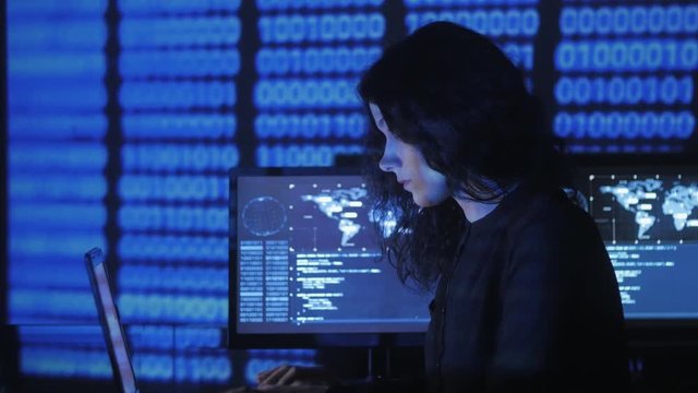portrait of young woman programmer working at a computer in the data center filled with display screens