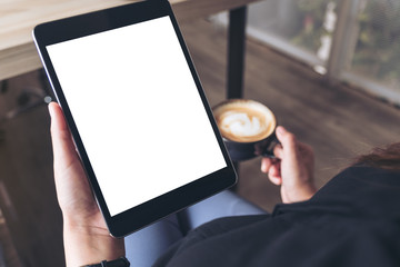 Mockup image of a woman sitting cross legged and holding black tablet pc with blank white desktop screen while drinking coffee in cafe