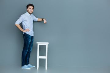 Useful object. Nice good looking handsome man standing near the stool and holding her hand above it while smiling to you