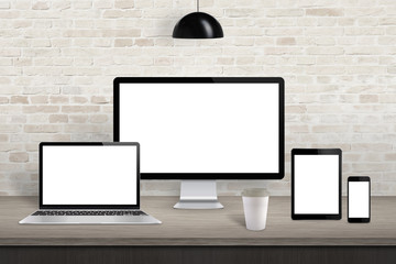 Fototapeta premium Computer display, laptop, tablet and mobile phone with isolated screen on office desk. White screen for web site design promotion. Brick wall in background.