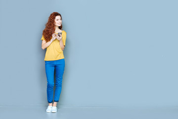 Digital device. Attractive cheerful young woman standing against blue background and smiling while using her mobile phone