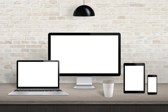 Computer display, laptop, tablet and mobile phone with isolated screen on office desk. White screen for web site design promotion. Brick wall in background.